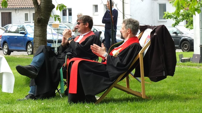 Zwei Personen in akademischen Roben klatschen und entspannen auf Liegestühlen im Freien während einer Veranstaltung.