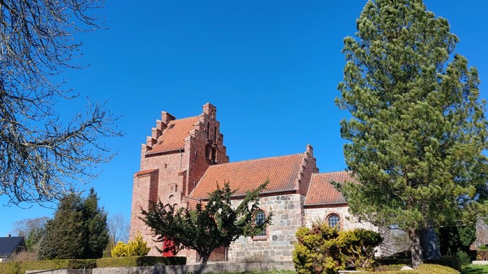 Historisk kirke med rødt tag og stenmure, omgivet af grønne områder og klar blå himmel