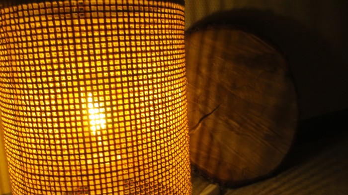 Eine zylindrische Lampe mit einem geflochtenen Schirm steht auf einem Tisch neben einem runden Kissen.