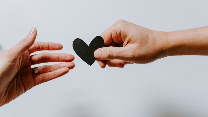 Zwei Hände tauschen ein kleines schwarzes herzförmiges Papier aus, das Zuneigung oder Liebe symbolisiert.
