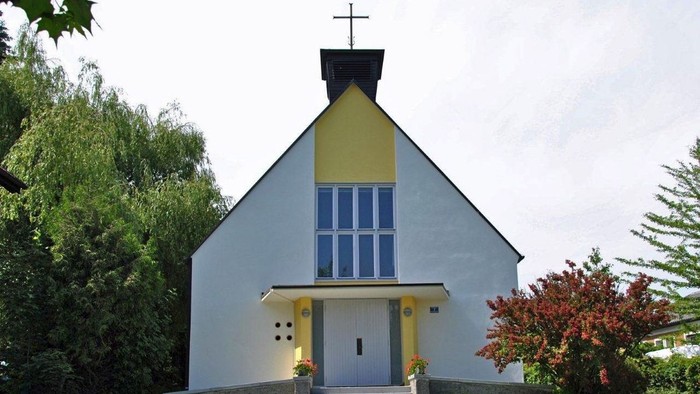 Kleine weiße Kirche mit gelben Akzenten und blauem Fenster, umgeben von Grün.