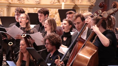 Ein Chor und ein Streichensemble treten gemeinsam in einem prunkvollen Konzertsaal auf und sind leidenschaftlich in ein Musikstück vertieft.