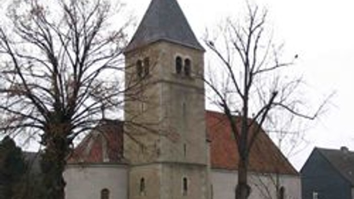 Kirchturm mit spitzem Dach und Uhr, umgeben von kahlen Bäumen und Häusern
