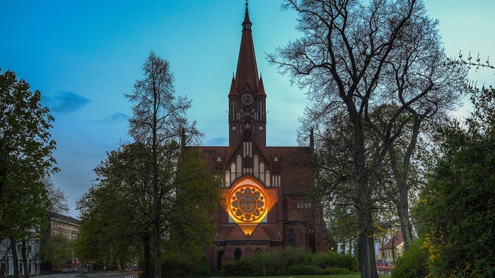 Historische Kirche, beleuchtet mit einem leuchtenden Rundfenster, umgeben von Bäumen und Park.
