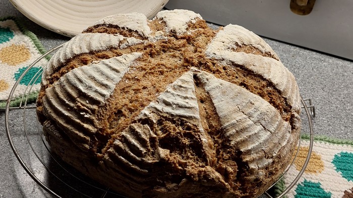 Frisch gebackenes Sauerteigbrot mit Kreuzschnitten auf einem Kühlgitter neben einer Backschüssel und einem Ofen.