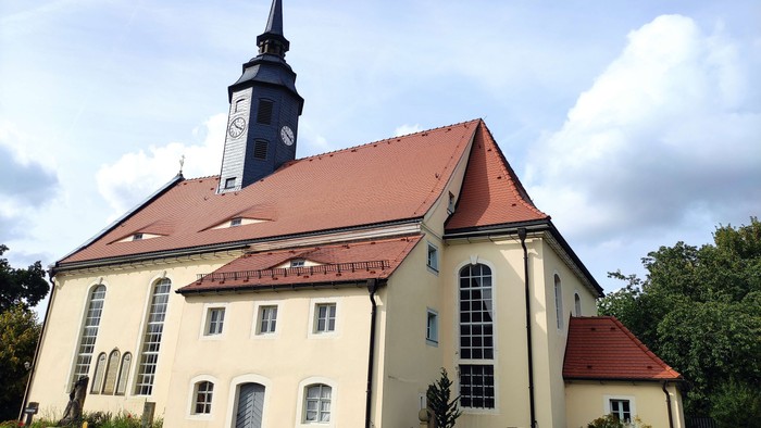 Ein weißes Kirchengebäude mit rotem Dach und hohem Turm, umgeben von Bäumen und einem Friedhof.