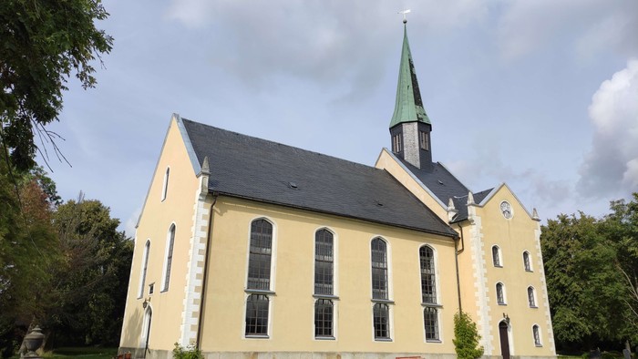 Gelbe Kirche mit hohem Turm und grünem Dach, umgeben von Bäumen und Gräbern