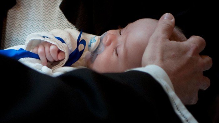 En baby sover på en persons brystkasse, iført en blå og hvid bodystocking.