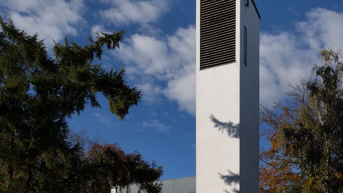 Moderne Kirche mit hohem weißem Turm und Kreuz an der Spitze, umgeben von Bäumen.