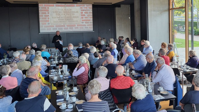 En stor gruppe af ældre mennesker sidder ved bord i et lokalet og spiser.