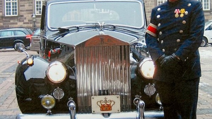 En uniformeret betjent står ved siden af en elegant, vintage Rolls-Royce foran en historisk bygning.