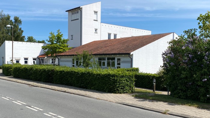 Et stort, kvadratisk hvidt hus med rødt tag og en højere sidefløj, beliggende ved en vej med buske og træer foran