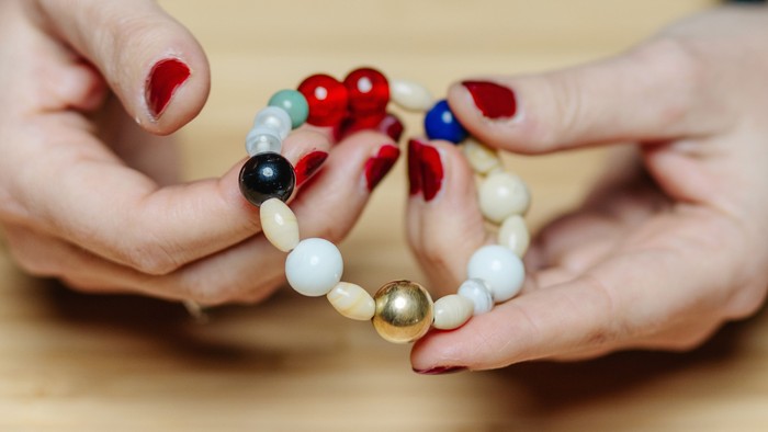 Hænder, der holder et farverigt perlearmbånd med røde, blå, sorte, hvide og gyldne perler.