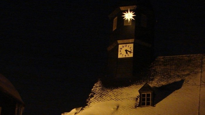 Schneebedeckte Häuser bei Nacht mit beleuchteter Kirchturmspitze
