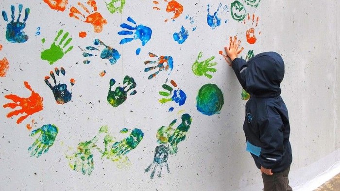 Ein Kind macht einen farbigen Handabdruck an eine Wand. Es sind schon viele bunte Handabdrücke auf der Wand