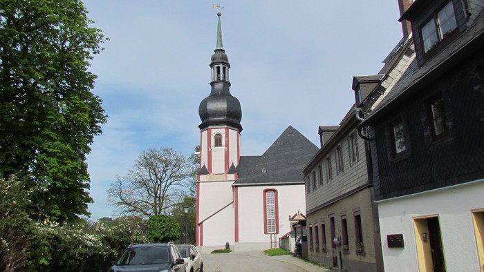 Kirchengemeinde mit hohem Turm und rot-weißer Fassade, umgeben von Bäumen und Häusern