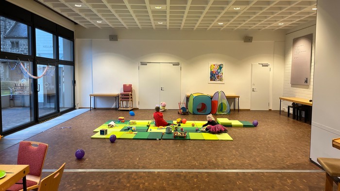 Ein großer Raum mit Kindern, die auf bunten Matten spielen, umgeben von Spielzeugen und Tischen