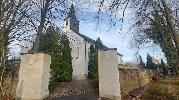 Kircheneingang mit zwei Säulen und Torbogen, umgeben von Bäumen und Mauern