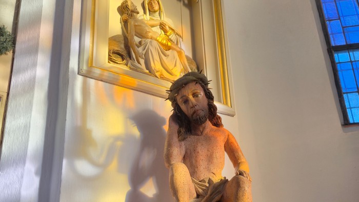 Eine Statue von Christus, der das Kreuz trägt, auf einem Sockel mit religiösen Kunstwerken darüber.