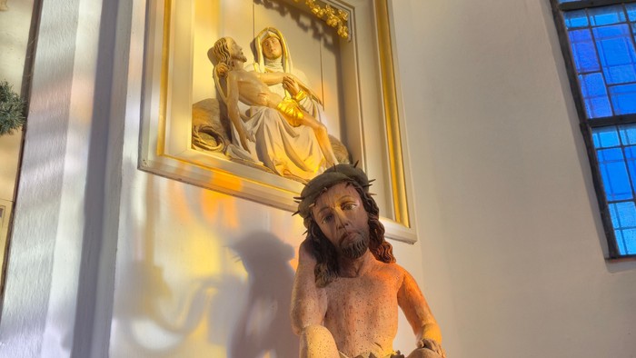 Eine Statue von Christus, der das Kreuz trägt, auf einem Sockel mit religiösen Kunstwerken darüber.