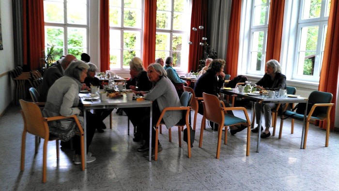 Eine Gruppe älterer Menschen sitzt in einem hellen Raum mit großen Fenstern und genießt eine Mahlzeit an Tischen.