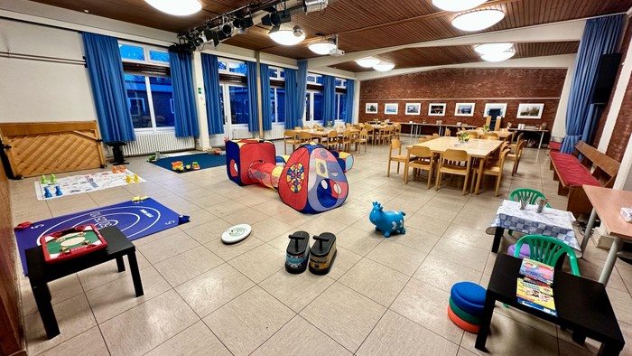 Ein großer Raum mit Kinder-Spielzeug, Tischen und Stühlen, blauen Vorhängen und Fenstern.