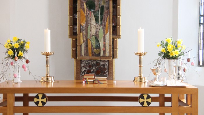 En altertavle med to lyse lysestake, to blomsterstråer og et maleri.