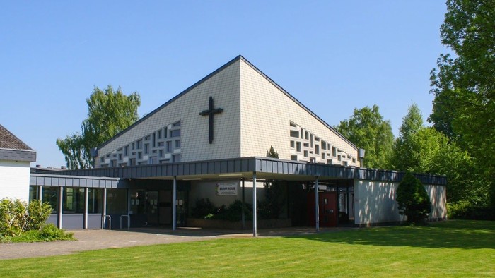 Das Bild zeigt eine moderne Kirche mit einem großen Kreuz an der Fassade, umgeben von Grünflächen und Bäumen.