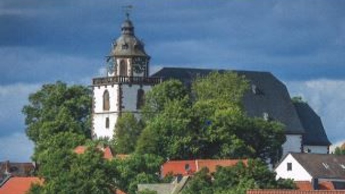 Eine große weiße Kirche mit einem hohen Turm steht zwischen Bäumen und rotbedachten Gebäuden unter einem blauen Himmel.