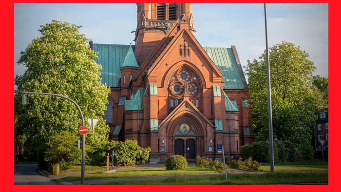 Das Bild zeigt eine rote Kirche mit grünen Dächern und einem Uhrturm, umgeben von Bäumen und Gras.