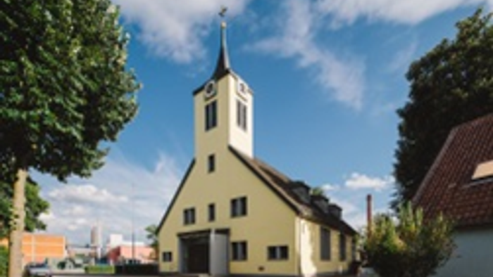 Eine gelbe Kirche mit einem hohen Turmspitz steht neben einem Gebäude mit rotem Dach unter einem blauen Himmel mit verstreuten Wolken.