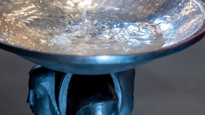 Eine Hand gießt Wasser in ein reflektierendes Metallbecken, das von einem dunklen skulpturalen Sockel getragen wird.