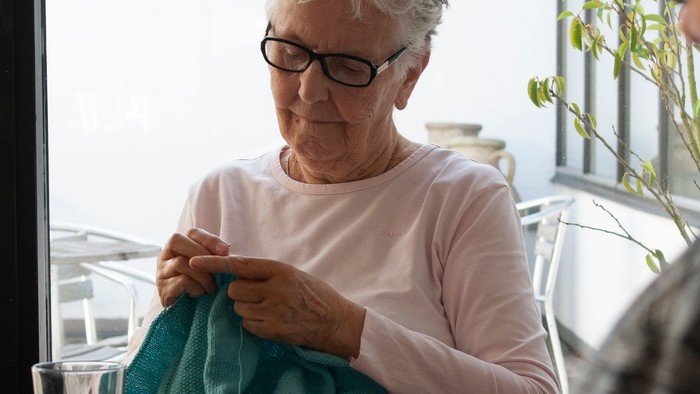 En ældre kvinde sitter ved vinduet og stråler en blå trøje.