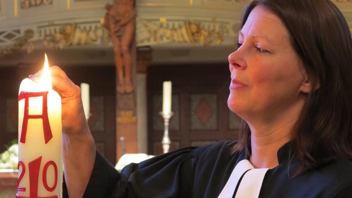 Frau in schwarzem Gewand hält eine brennende Kerze mit rotem Kreuz und Zahlen.