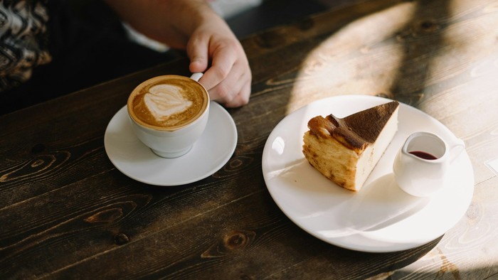 Eine Person hält eine Tasse Kaffee neben einem Stück Kuchen auf einem Holztisch