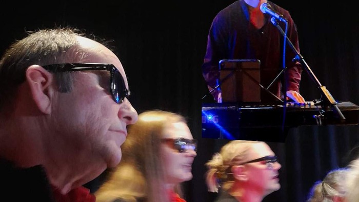 Et publikum ser en optrædende synge og spille keyboard på scenen med dramatisk belysning.