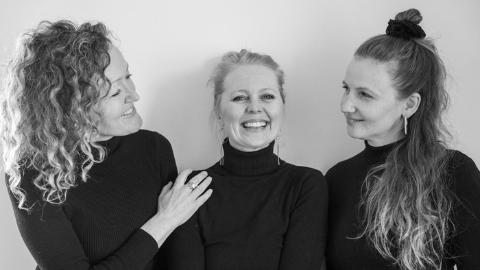 Tre glade kvinder i sorte toppe, der poser tæt sammen i et venligt gruppeportræt.