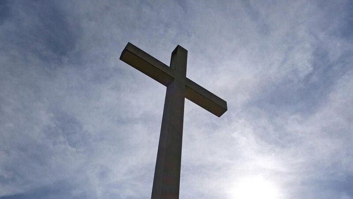 Ein hohes Kreuz ragt markant vor einem dramatischen, sonnenbeschienenen Himmel mit verstreuten Wolken auf.
