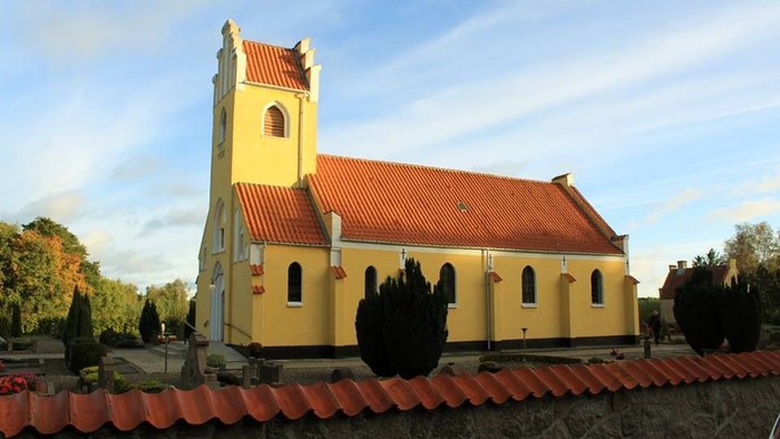 Gul kirke med rødt tag og tårn, omgivet af træer og mur.