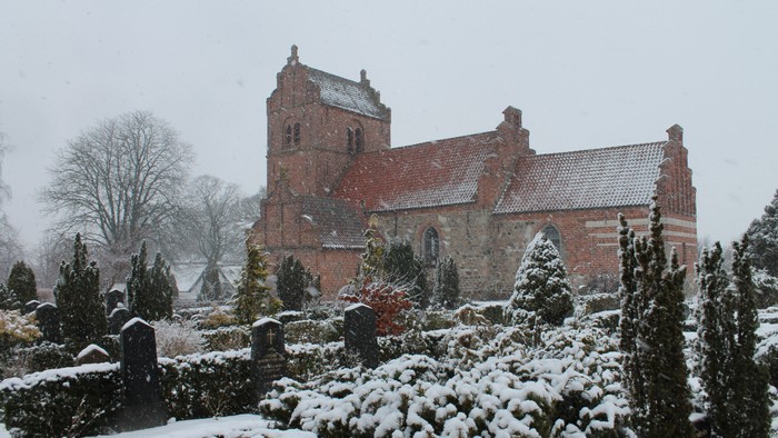 En snedækket kirke med gravhøje og træer i forgrunden