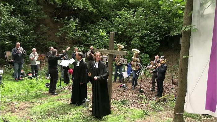 Freiluft-Trauerfeier mit Musikern, die Blechblasinstrumente spielen, und Geistlichen, die den Gottesdienst nahe einem Kreuz leiten.