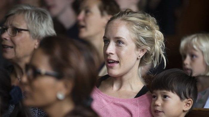 En kvinde i en lyserød skjorte holder et lille barn, mens hun deltager i en begivenhed med andre mennesker.