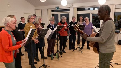 En gruppe mennesker, der synger sammen i et kor, ledet af en dirigent med noder.