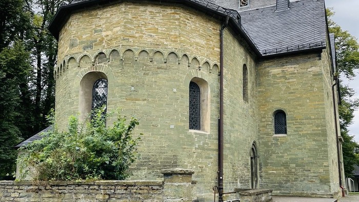 Alte Kirche mit spitzen Dach und kleinen Fenstern, umgeben von Bäumen und einem Steinmauer.