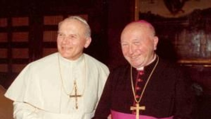 Zwei Männer in religiöser Kleidung, einer hält ein Buch, lächeln.