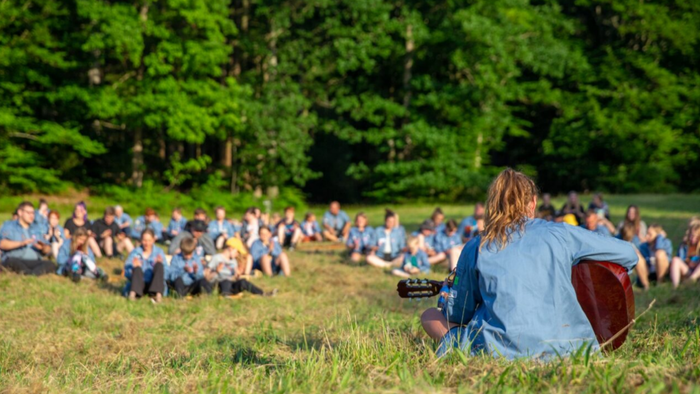 En gruppe mennesker sidder på græsset og kigger mod en scene i en park.