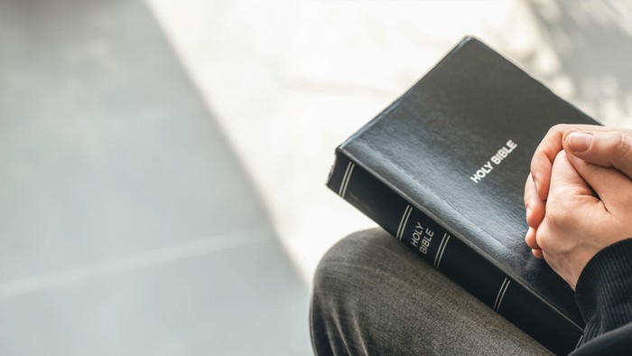person sitting with their hands folded on a bible placed on their knees