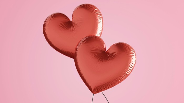 Zwei glänzende rote herzförmige Ballons, die zusammen vor einem zarten rosa Hintergrund schweben.