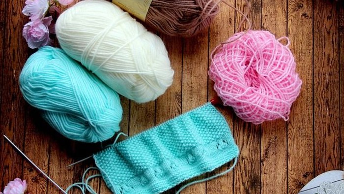 Farverige garnnøgler, strikkepinde, en saks og en færdig strikket lyseblå mundbind på en træoverflade.