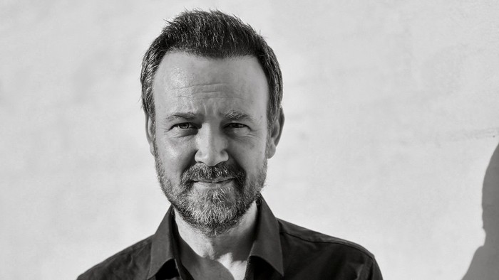 En mand med skæg i en knap-op skjorte poser mod en ensfarvet baggrund.